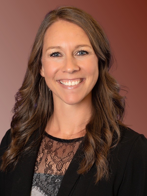 A brunette woman wearing a black blazer smiles at the camera in front of a maroon background