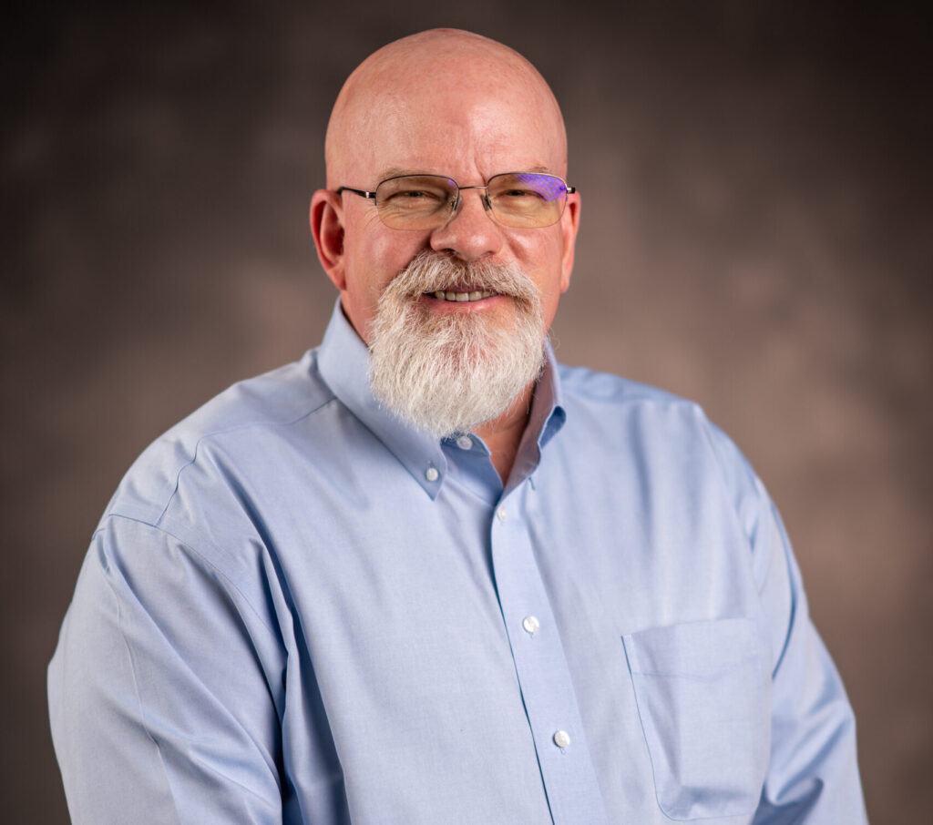 Bald man in blue button-down shirt, with a full gray beard, smiles at the camera.