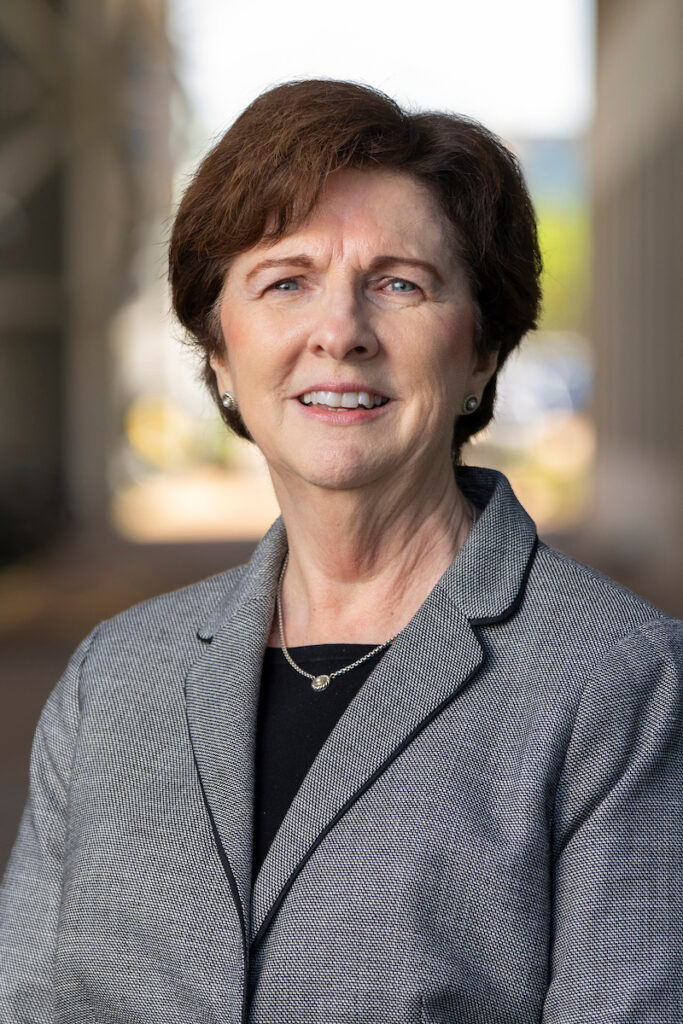 A woman with short-cropped brown hair wears a black shirt and gray blazer, and smiles at the camera.