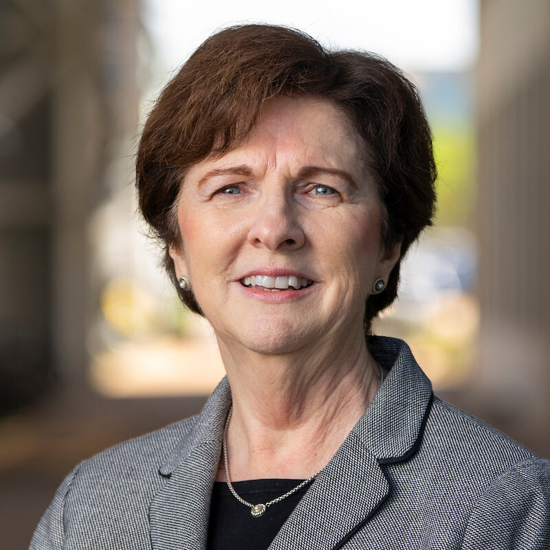 A woman with short-cropped brown hair wears a black shirt and gray blazer, and smiles at the camera.