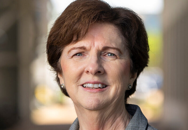 A woman with short-cropped brown hair wears a black shirt and gray blazer, and smiles at the camera.