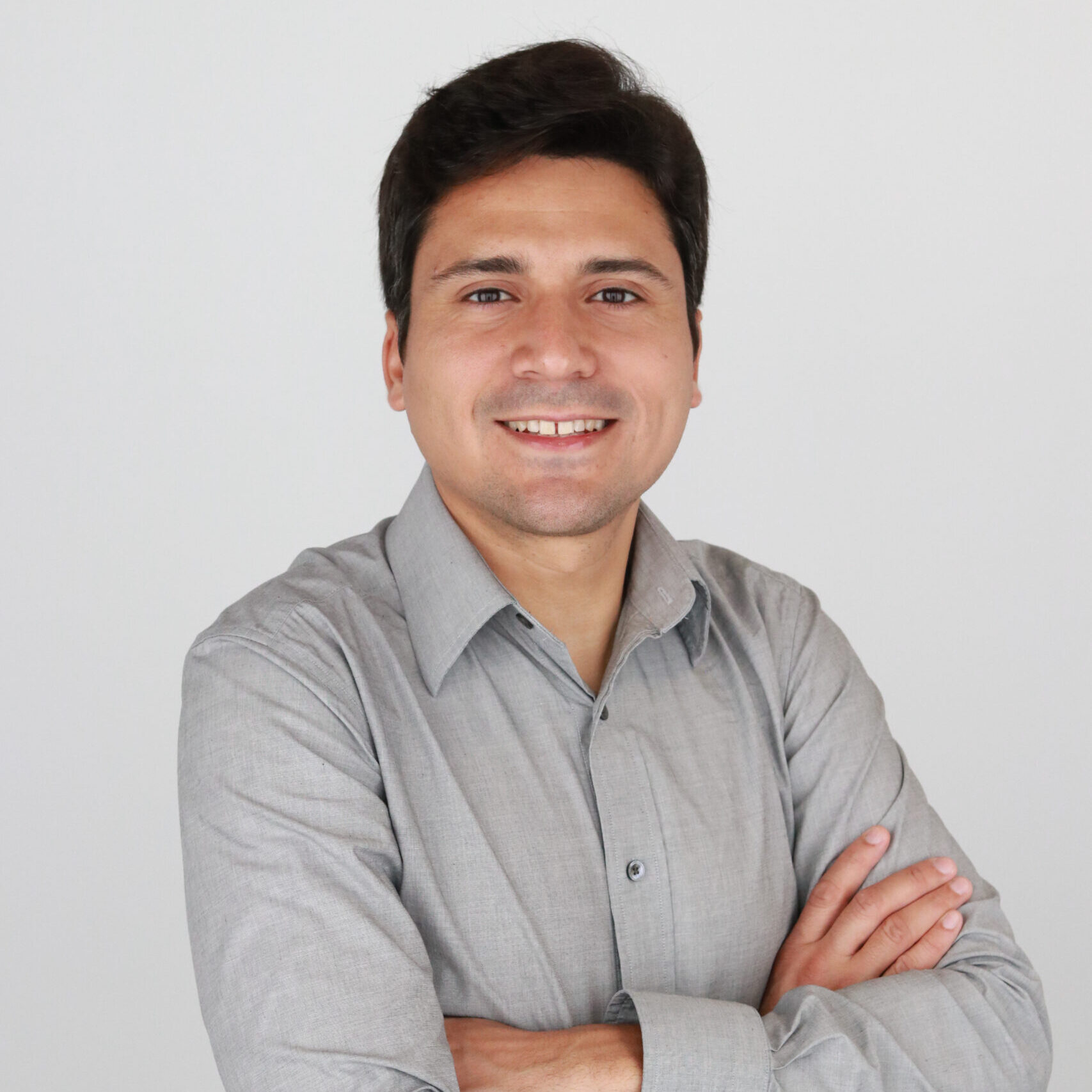 Man with close-cut dark hair wears a gray button-down shirt, crosses arms over chest and smiles at camera