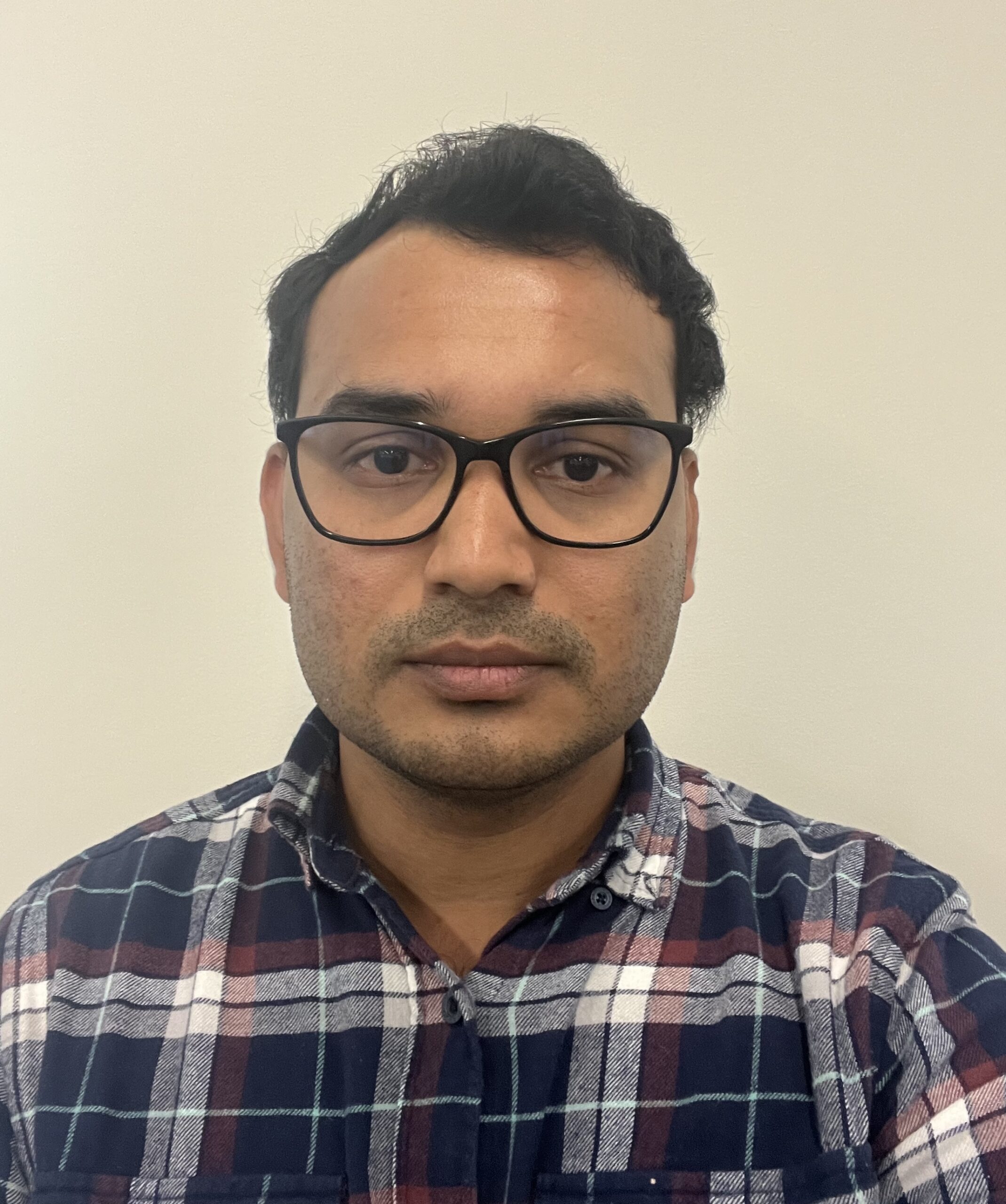 Man with dark hair and glasses wears a plaid shirt and looks into the camera